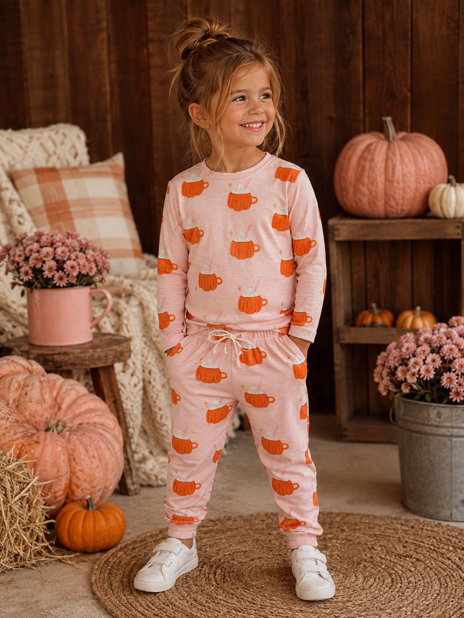 Smiling girl in cozy pink pumpkin-patterned outfit, standing among pumpkins and flowers, in a warm, autumn setting.