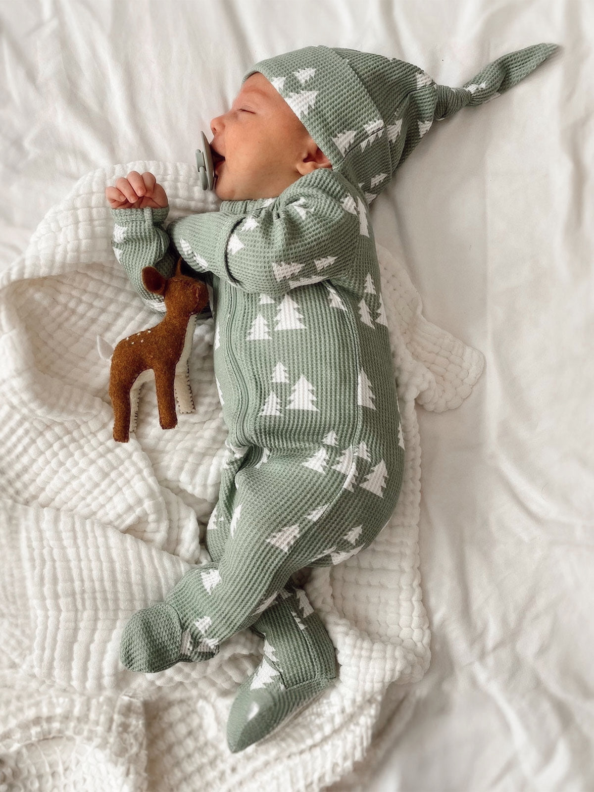 Baby sleeping on a soft blanket, wearing a green patterned outfit and hat, holding a toy deer.