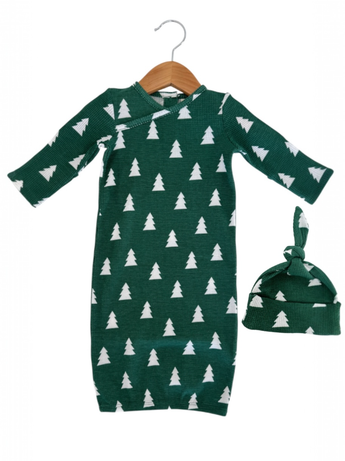 Green baby gown with white tree patterns, paired with a matching knotted hat, hanging on a wooden hanger.