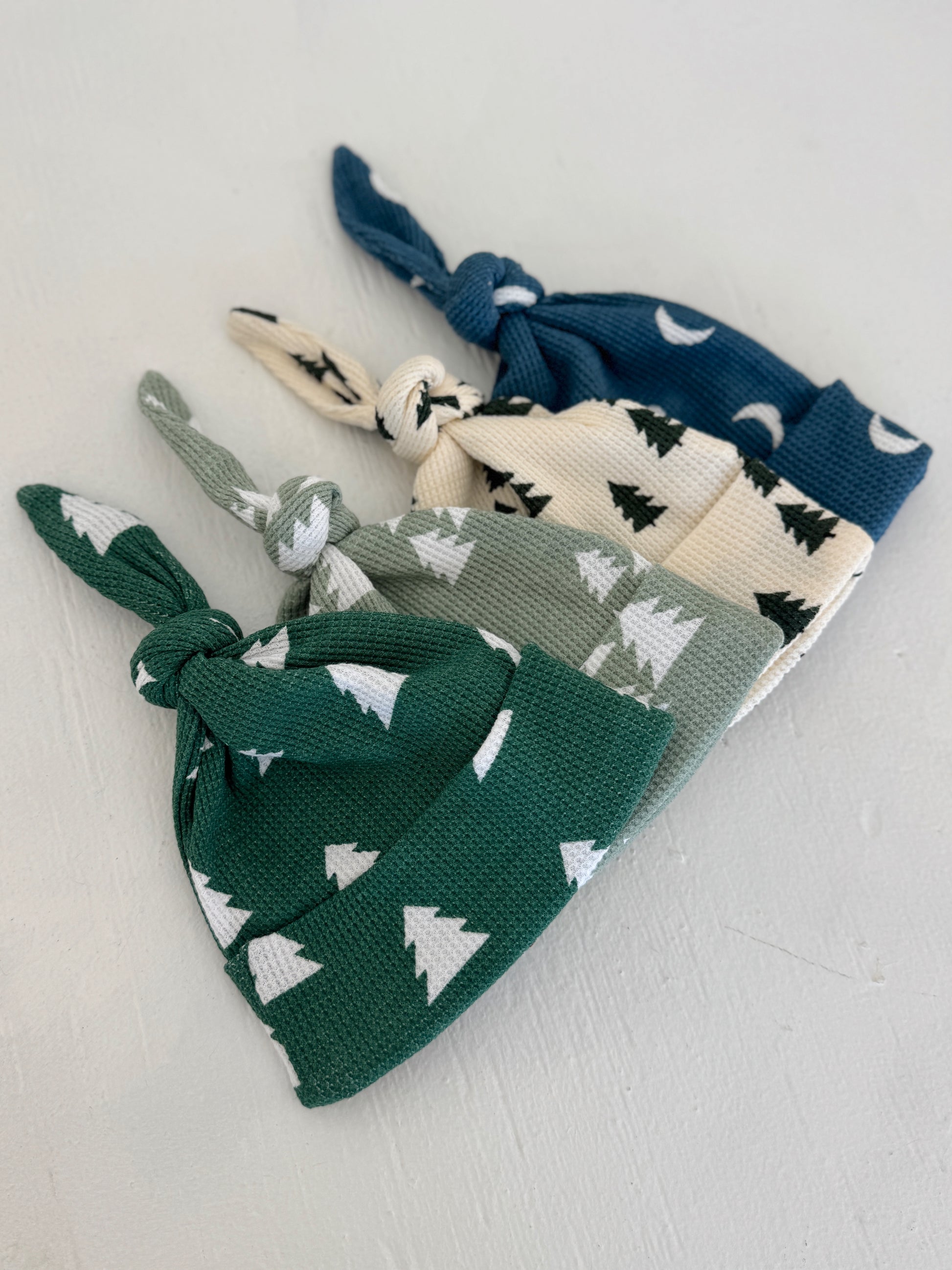 Four cozy knotted hats in green, cream, and blue with tree and moon patterns arranged neatly on a light background.