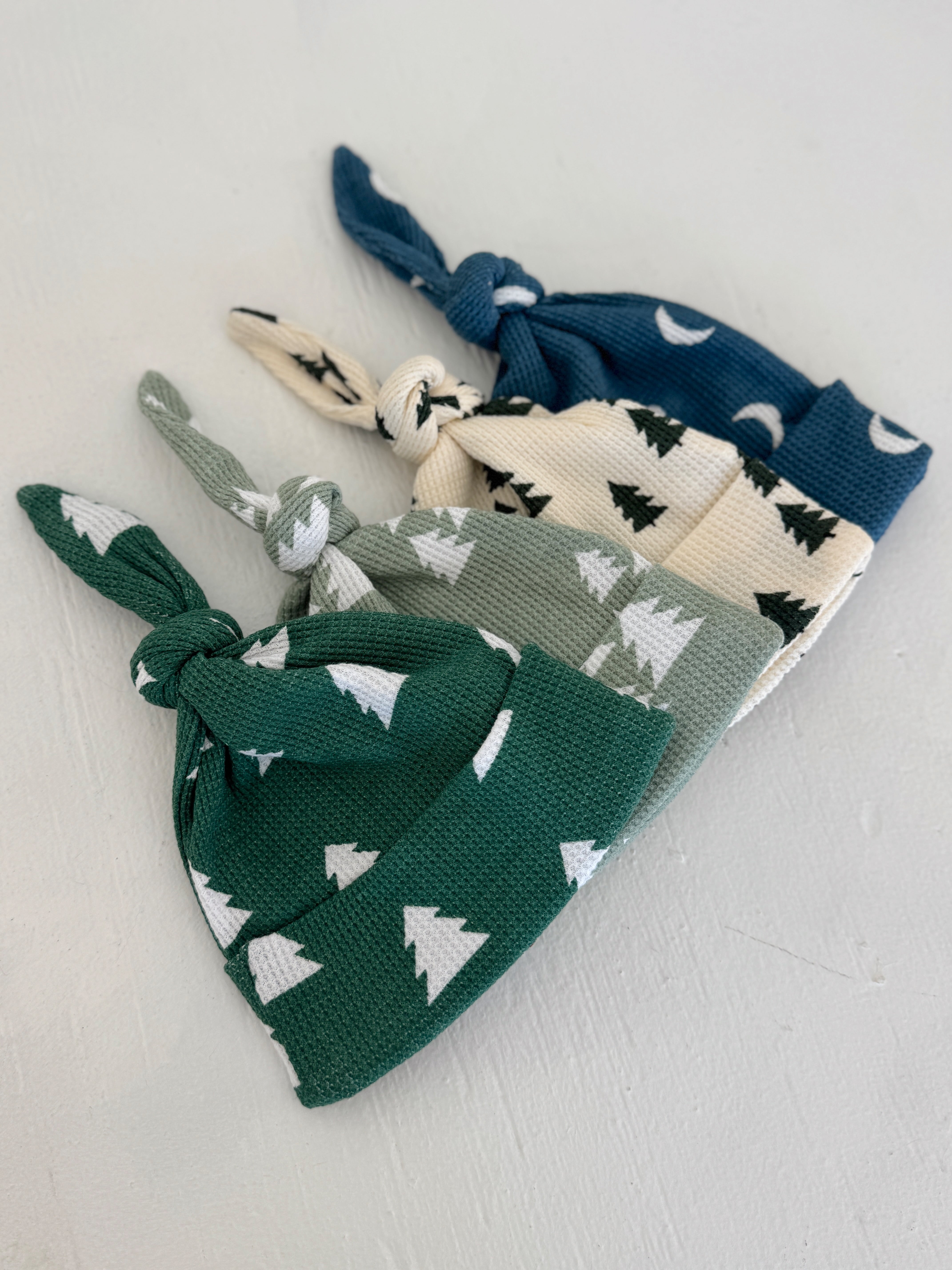 Four stylish baby knot hats in green, blue, cream, and gray with tree patterns on a light background.