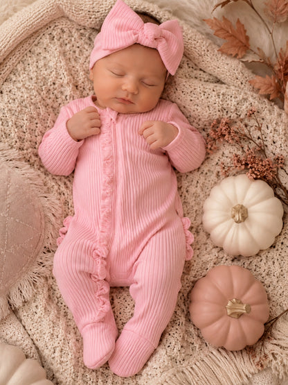 Newborn baby girl in pink outfit and bow, lying on a soft blanket with decorative pumpkins and autumn leaves.