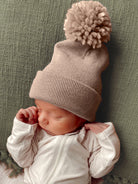 Newborn wearing a cozy beige knit hat with a pom-pom, resting on a textured green background.