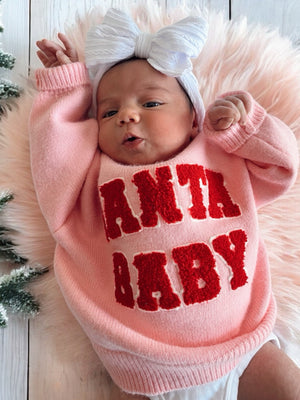 A baby in a pink sweater with "SANTA BABY" text and a white headband, lying on a fluffy pink surface.