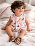 Smiling toddler with curly hair sits on a soft blanket, wearing a playful outfit with food patterns and sandals.