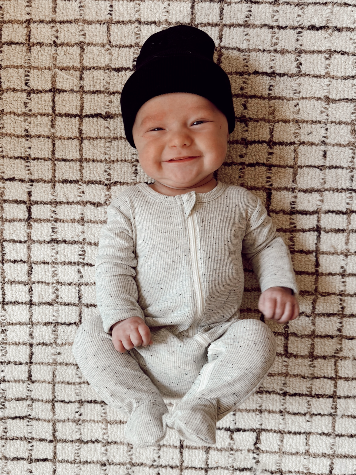 Smiling baby in a cozy, light-colored onesie and black beanie, sitting on a textured, patterned carpet.