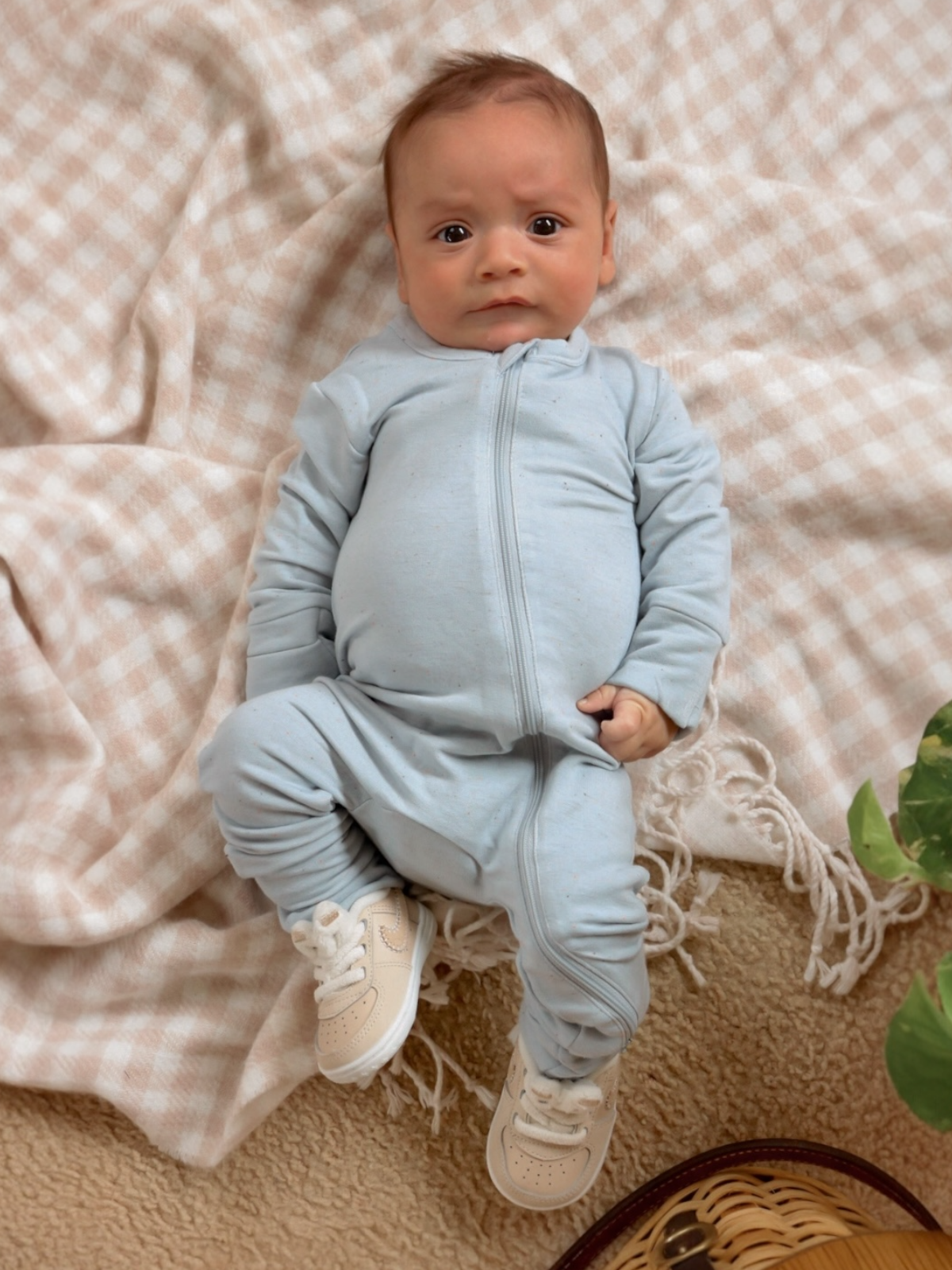 Infant in light blue outfit sits on a soft blanket, wearing shoes, with a curious expression.