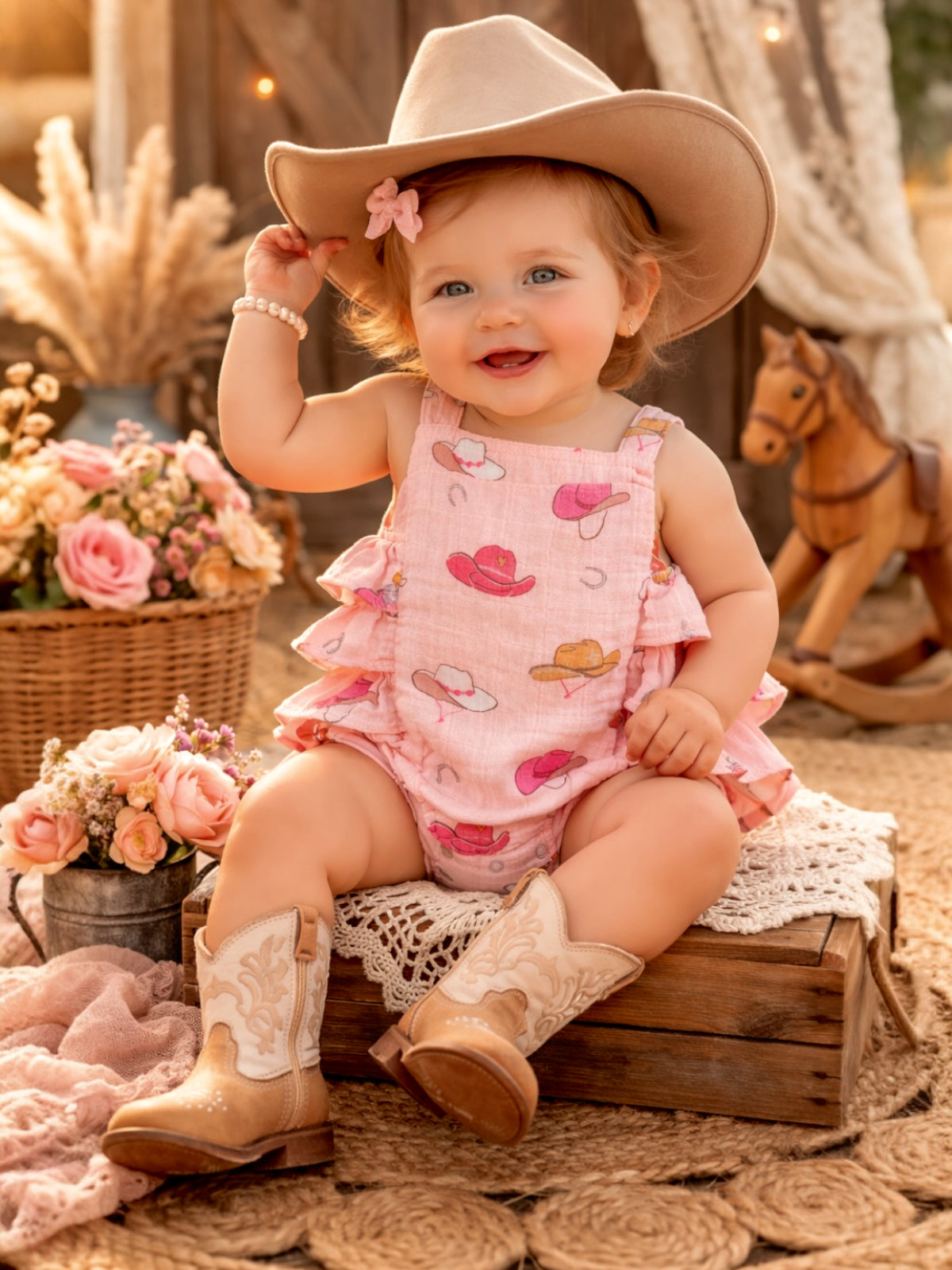 Smiling baby girl in a pink hat-patterned outfit and cowboy boots, wearing a hat and surrounded by flowers and decor.