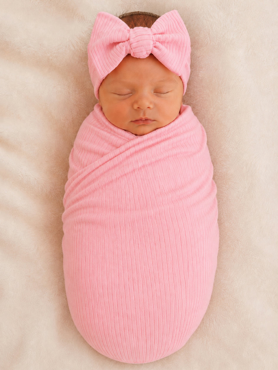 Newborn baby swaddled in a pink fabric with a matching bow headband, resting on a soft surface.