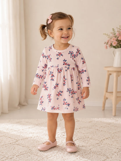 Smiling toddler in a floral dress and pink shoes, standing on a rug with soft lighting and a vase of flowers nearby.