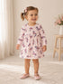 Smiling toddler in a floral dress and pink shoes, standing on a rug with soft lighting and a vase of flowers nearby.