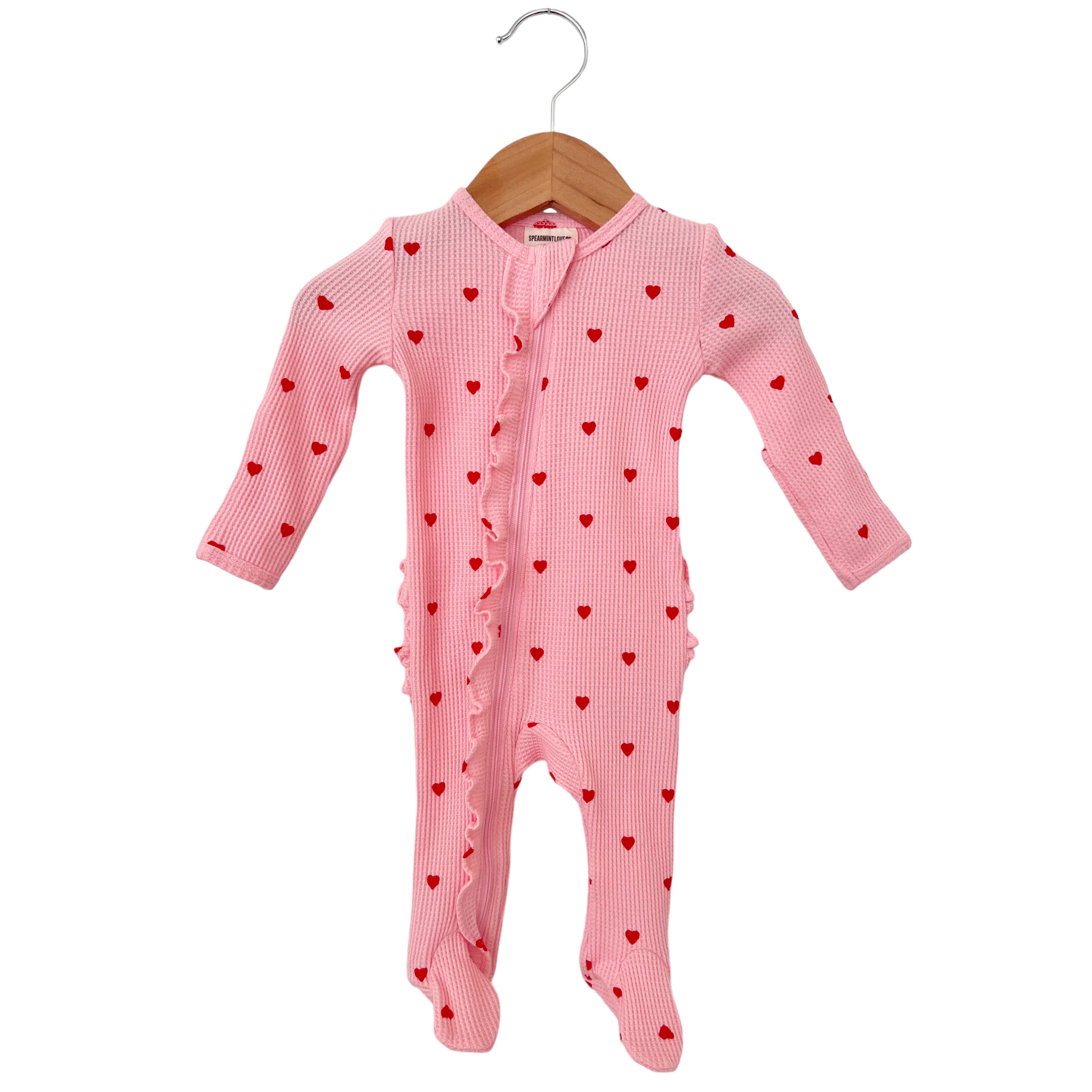 Pink baby onesie with red heart prints and ruffle detailing, hanging on a wooden hanger.