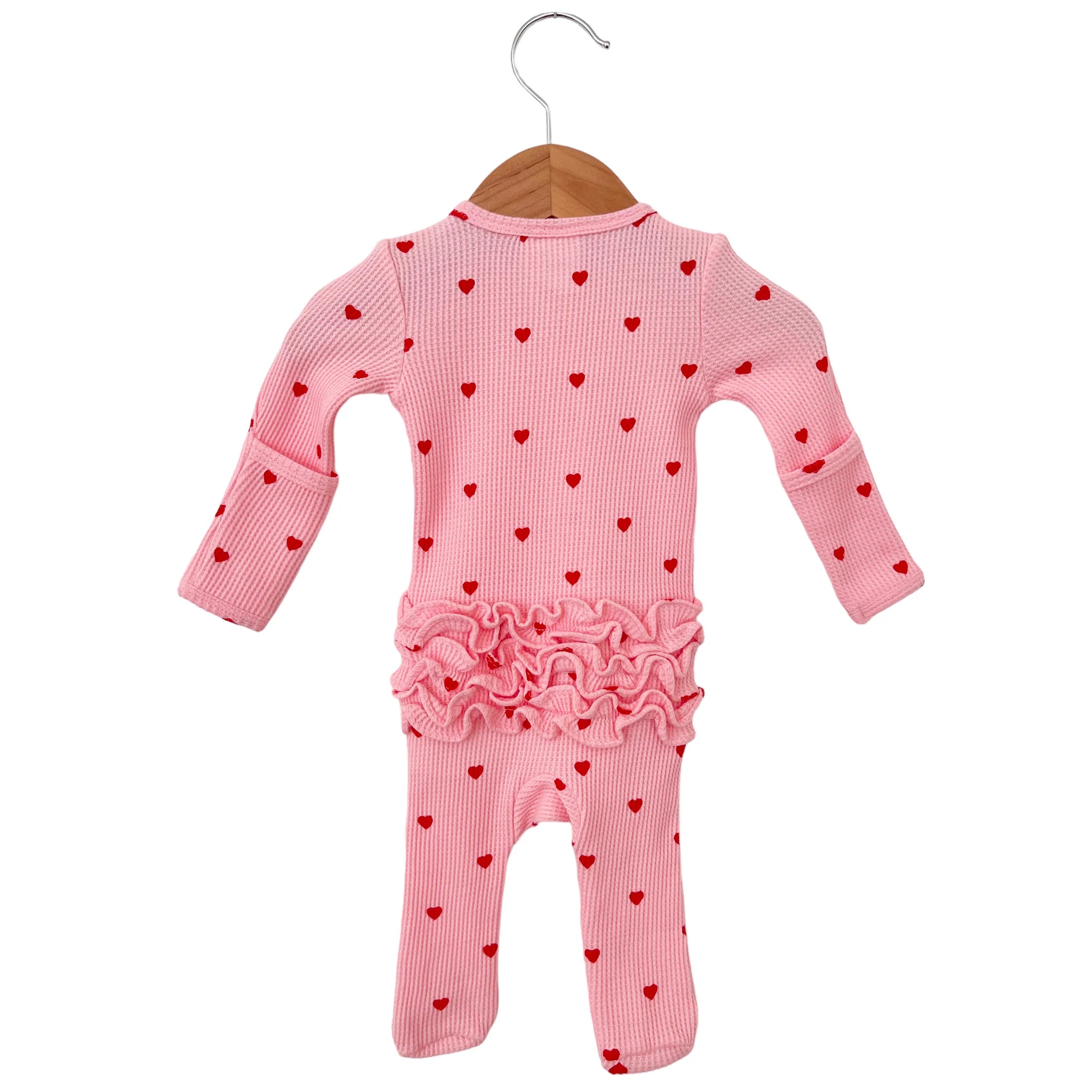 Pink baby onesie with red heart patterns and ruffled hem, hung on a wooden hanger.