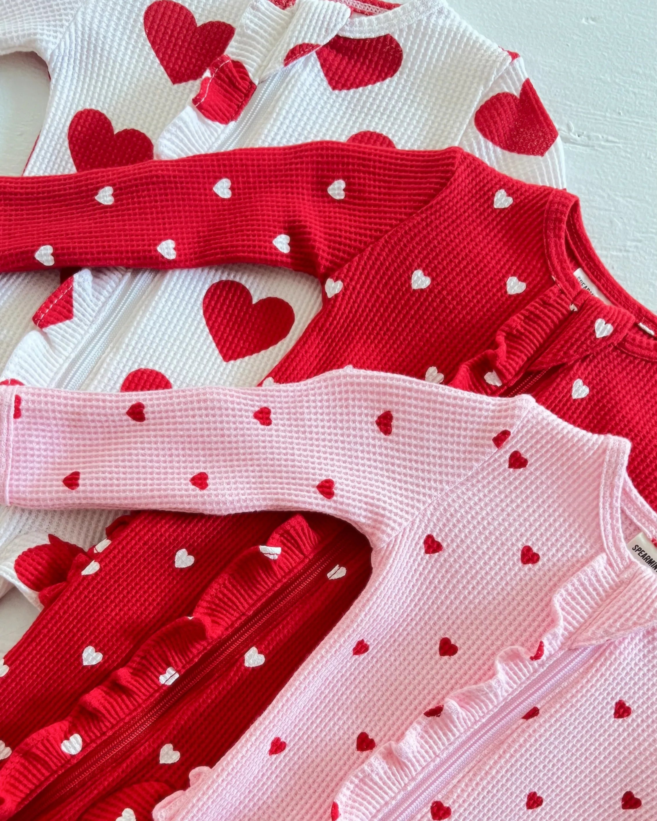 Three knitted cardigans in red, white, and pink, adorned with small heart patterns.