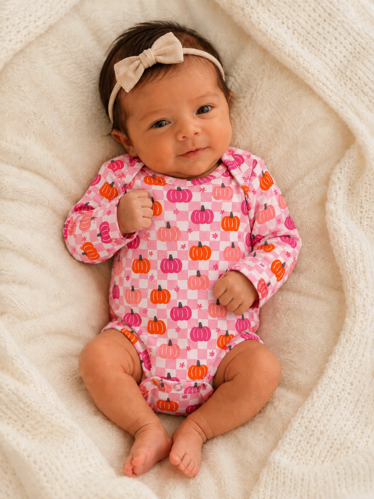 Smiling baby in a pink pumpkin-patterned onesie, laying on a soft, cream-colored blanket.