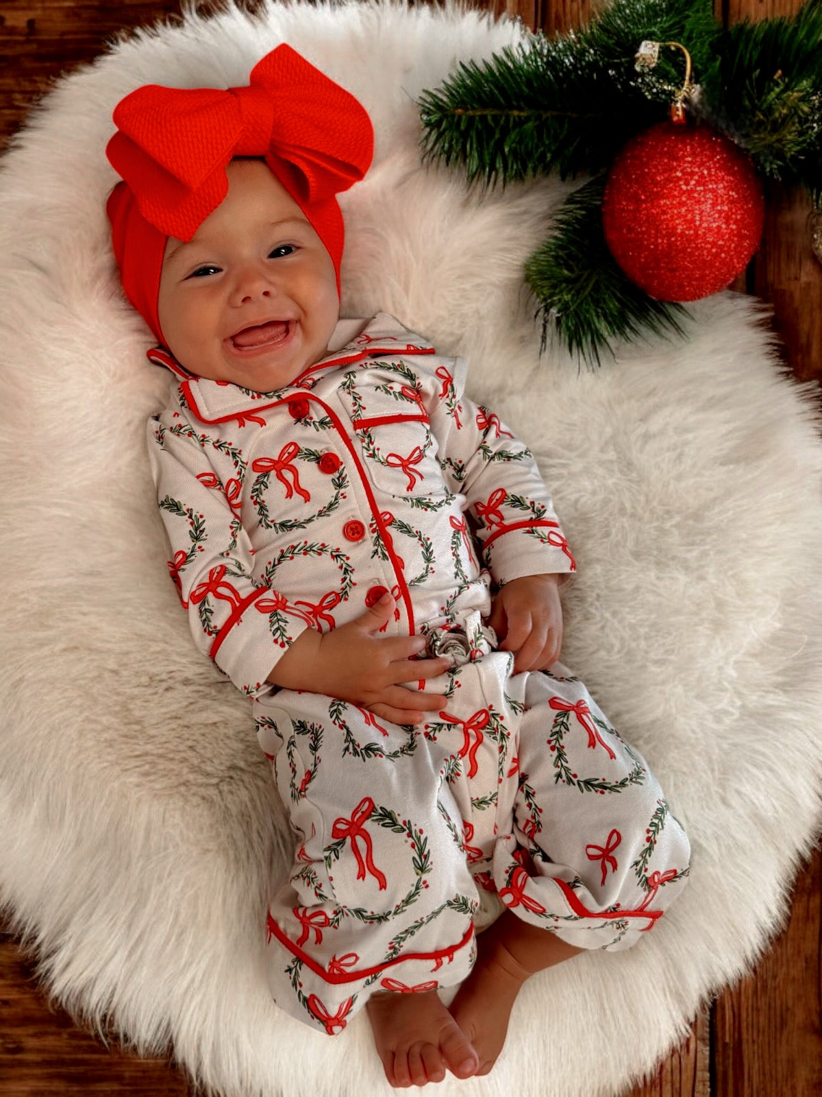 Smiling baby in festive outfit with red bow, surrounded by holiday decorations on a cozy white blanket.