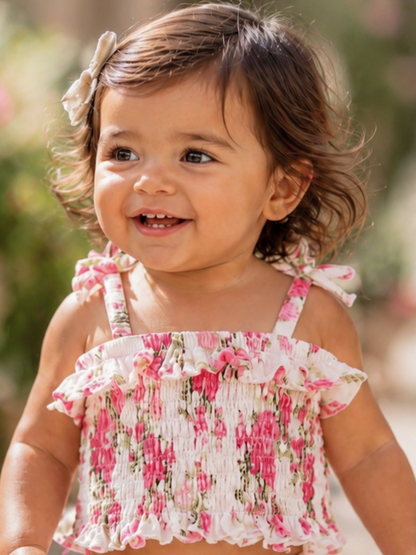 Smiling toddler with curly hair wearing a floral smocked top, enjoying a sunny day outdoors.