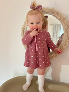 Smiling toddler in a heart-patterned dress holding a pink toothbrush, standing by a decorative mirror.