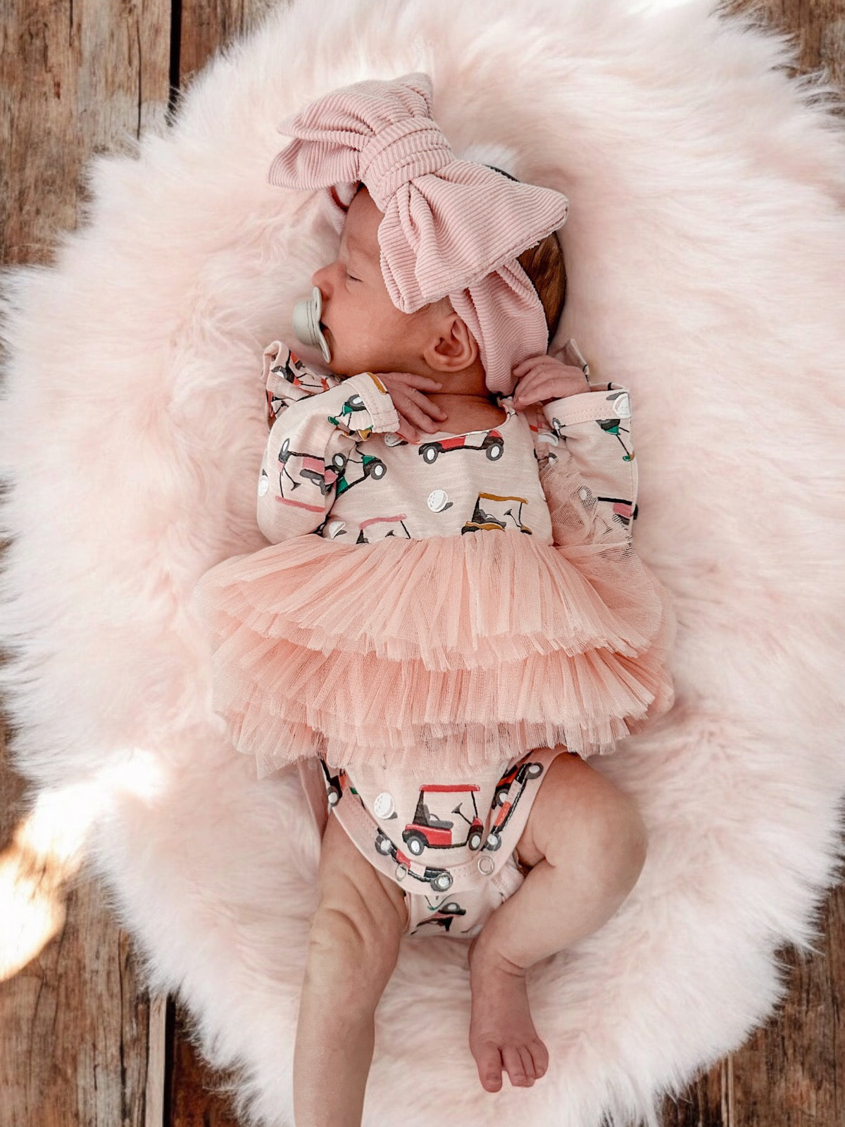 Cute baby girl wearing a pink bow and a dress with cars, lying on a fluffy rug.