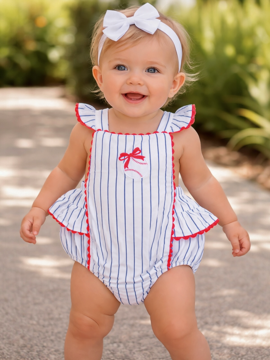 Smiling baby girl in a striped swimsuit with a bow, standing outdoors on a sunny day.