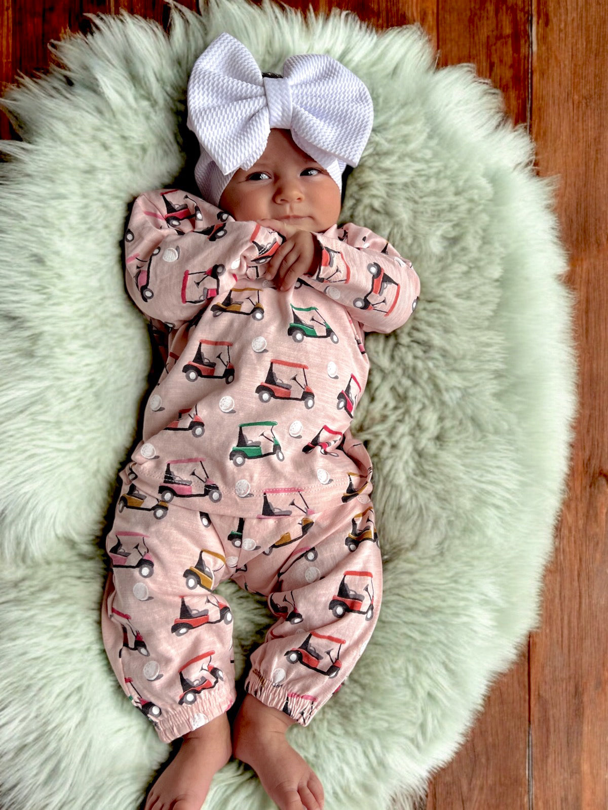 Baby in a pink golf cart print outfit with a large white bow on a fluffy green rug.