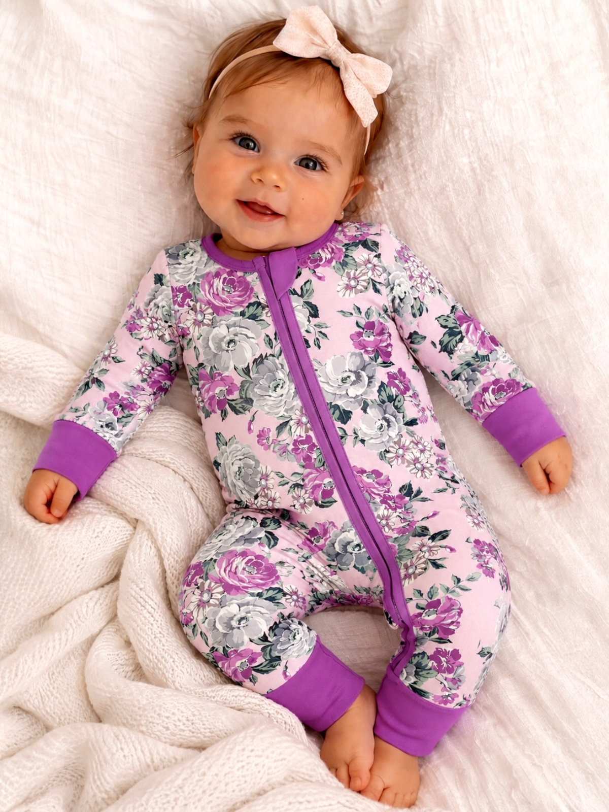 Smiling baby in floral-patterned pajamas with purple accents, sitting on a cozy white blanket.
