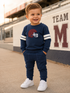 Smiling toddler in navy outfit with football design, standing on pavement near stadium, wearing white sneakers.