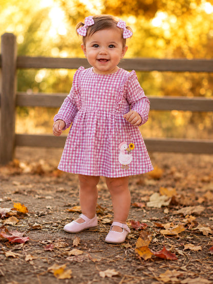 Smiling toddler in a pink gingham dress with bows, standing among autumn leaves near a wooden fence.