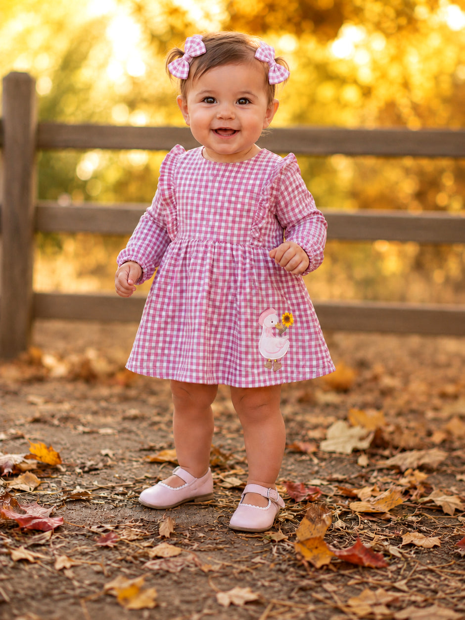 Smiling toddler in a pink gingham dress with bows, standing among autumn leaves near a wooden fence.