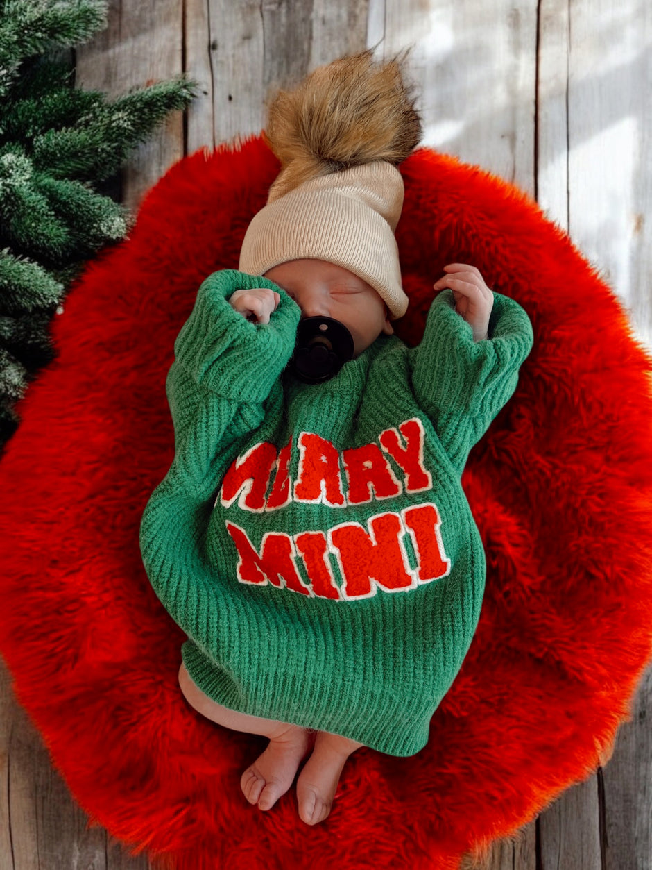 Newborn in a green sweater reading “Merry Mini,” resting on a red fuzzy blanket with a playful tan hat.