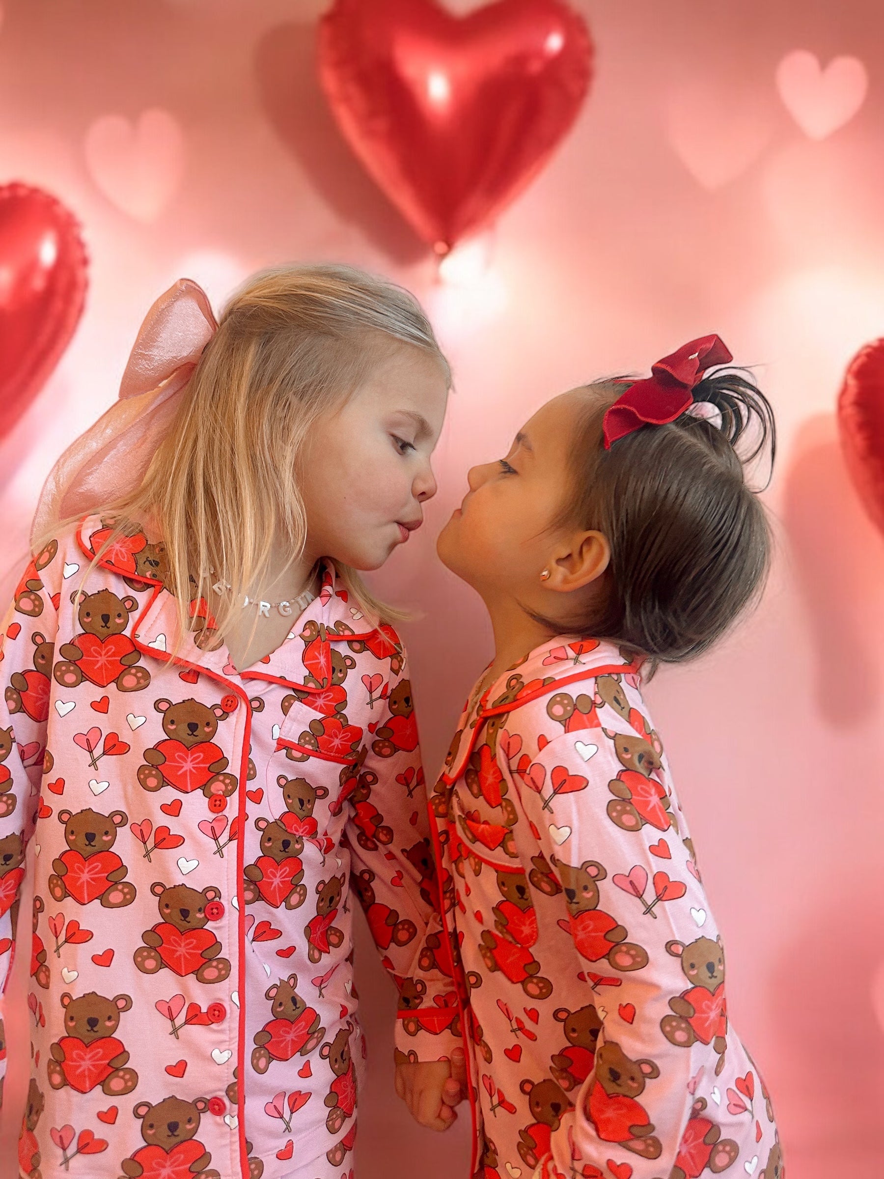 Two young girls in matching bear print pajamas kiss, surrounded by heart-shaped balloons on a pink backdrop.