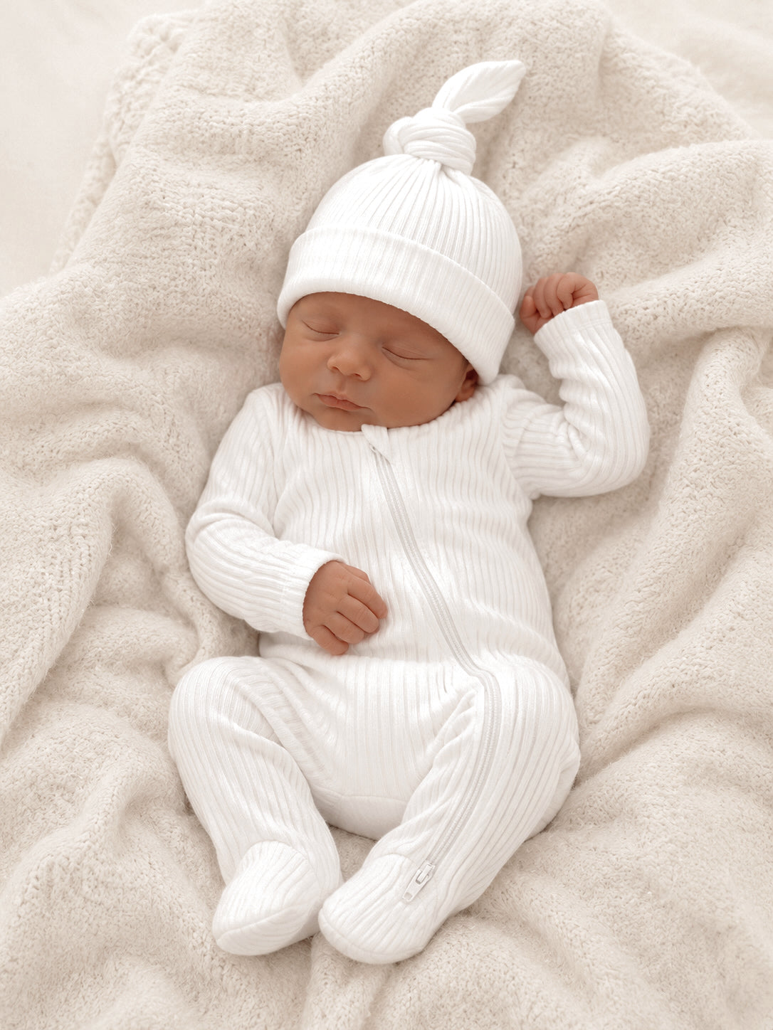 Sleeping baby dressed in a white ribbed outfit and hat, resting on a cozy light blanket.