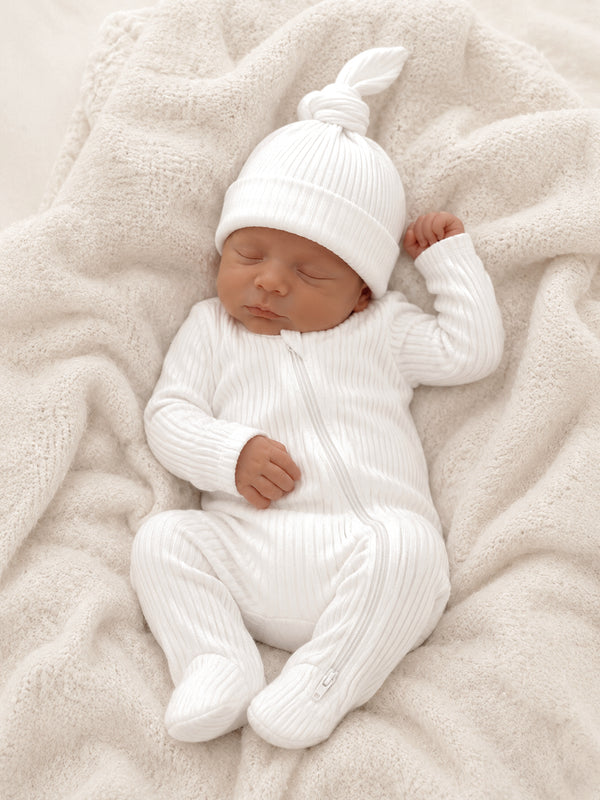 Sleeping baby dressed in a white ribbed outfit and hat, resting on a cozy light blanket.