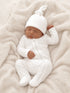 Sleeping baby dressed in a white ribbed outfit and hat, resting on a cozy light blanket.