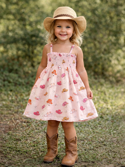 Smiling child in a patterned dress and boots, wearing a straw hat, standing outdoors among greenery.