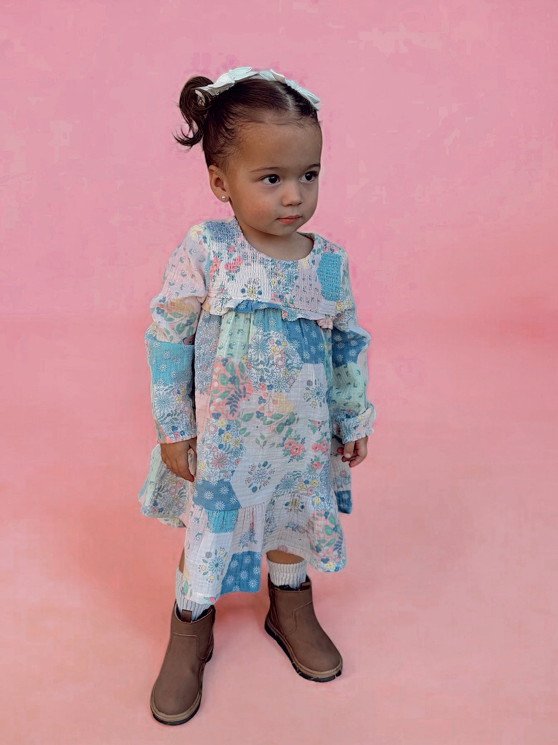 A young child in a colorful patchwork dress and brown boots stands against a pink background.