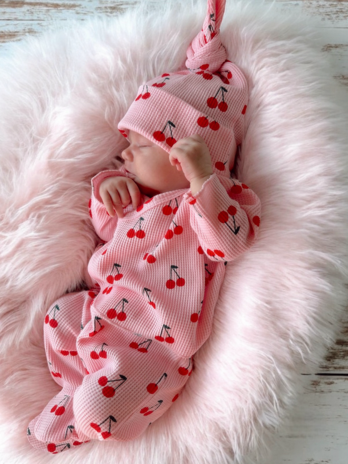 Newborn baby in cherry-patterned outfit and hat, resting on a fluffy pink surface.