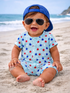 Smiling baby in star-patterned romper, wearing sunglasses and a blue cap, playing on a sandy beach.