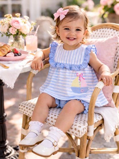 Smiling toddler in a striped dress sits in a chair, surrounded by flowers and a breakfast table with pastries.