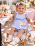 Smiling toddler in a striped dress sits in a chair, surrounded by flowers and a breakfast table with pastries.