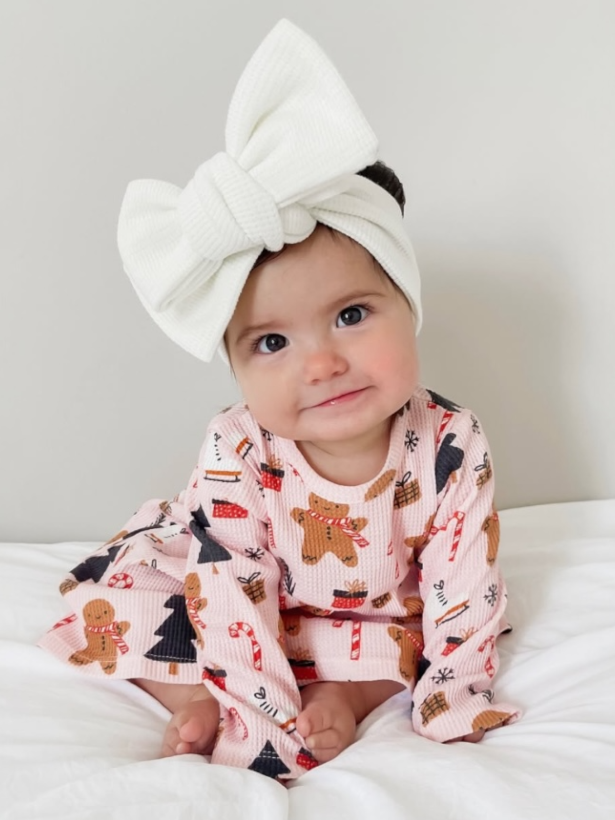 Smiling baby girl in a festive outfit and large white bow, sitting on a white blanket.