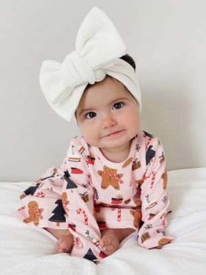 Smiling baby girl in a festive outfit and large white bow, sitting on a white blanket.