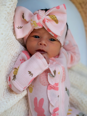 Infant wearing a pink floral onesie and large bow headband, resting on a cozy blanket.