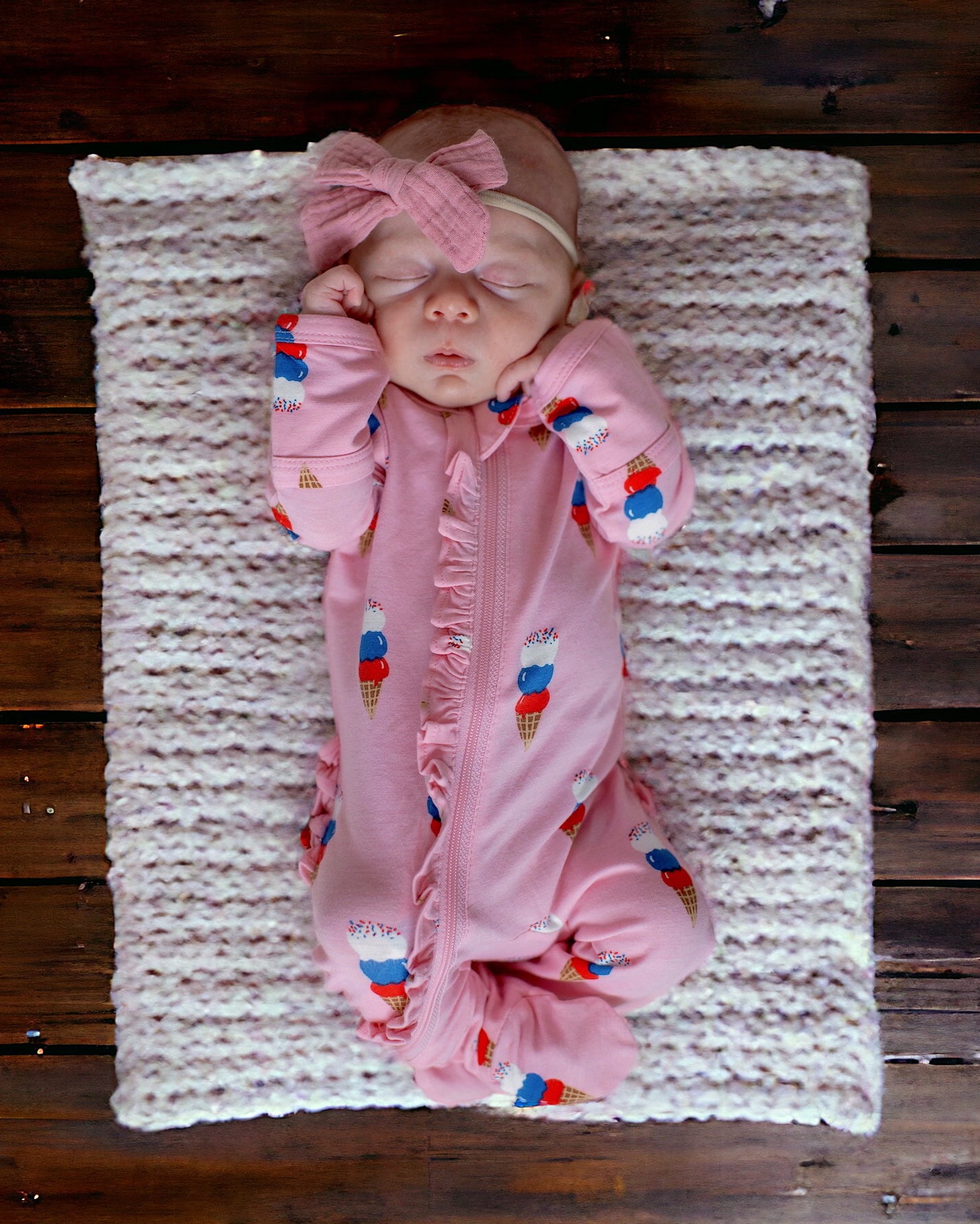 Sleeping baby in pink ice cream-themed outfit, resting on a soft blanket on wooden floor.