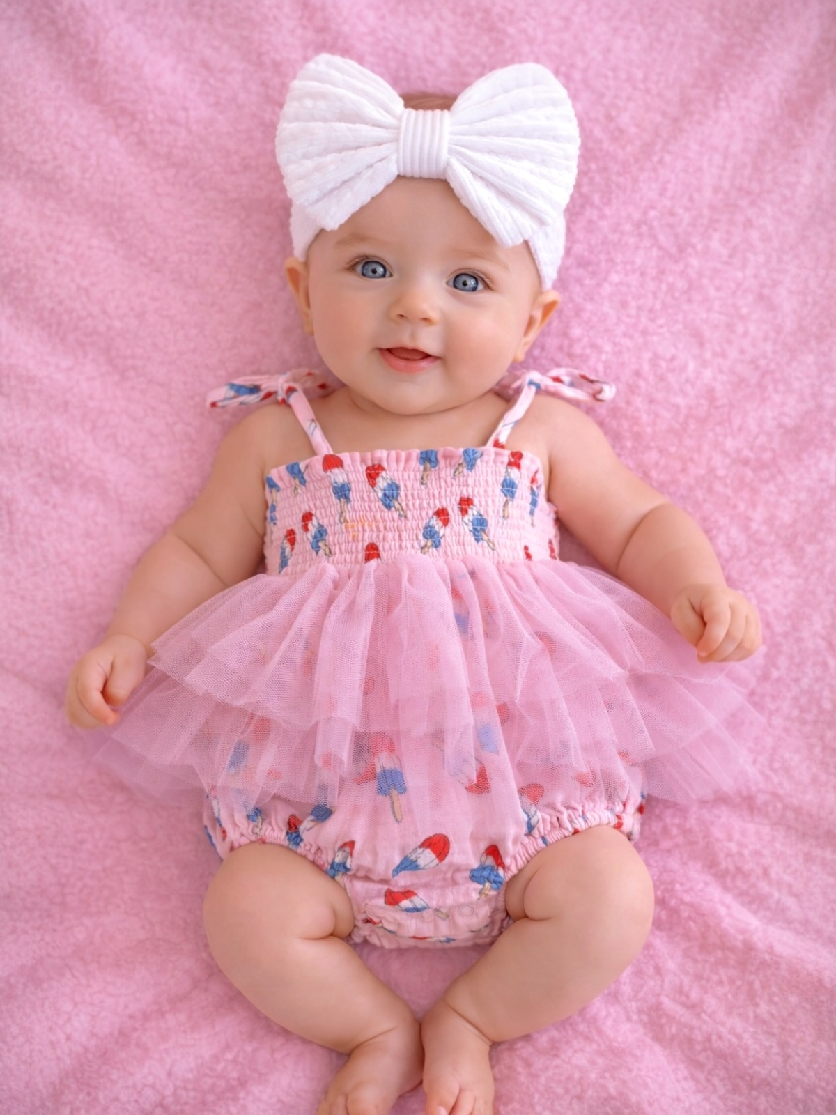 Baby in a pink smocked outfit with a tulle skirt and large white bow, smiling on a pink blanket.