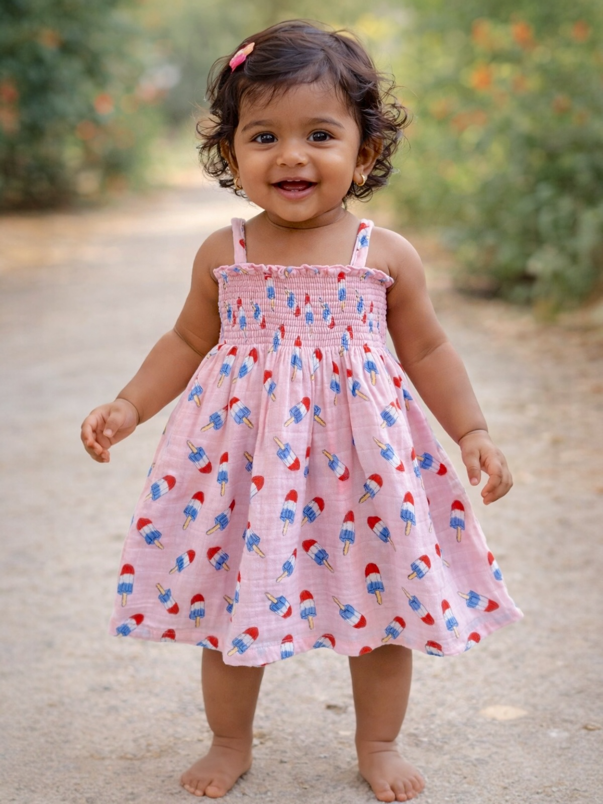 Smiling young child in a pink dress with popsicle print, standing on a path surrounded by greenery.