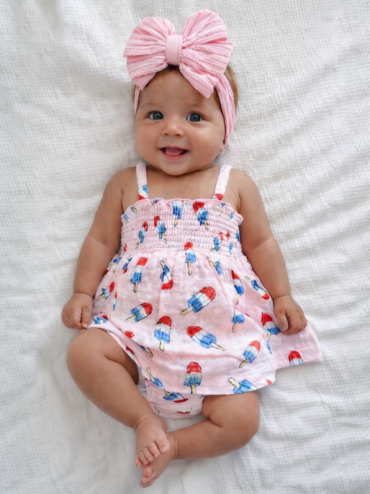 Smiling baby in pink dress with popsicle print and large pink bow, lying on a white textured blanket.