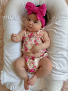 Baby girl with a pink bow, wearing a floral outfit, lying on a soft pillow.
