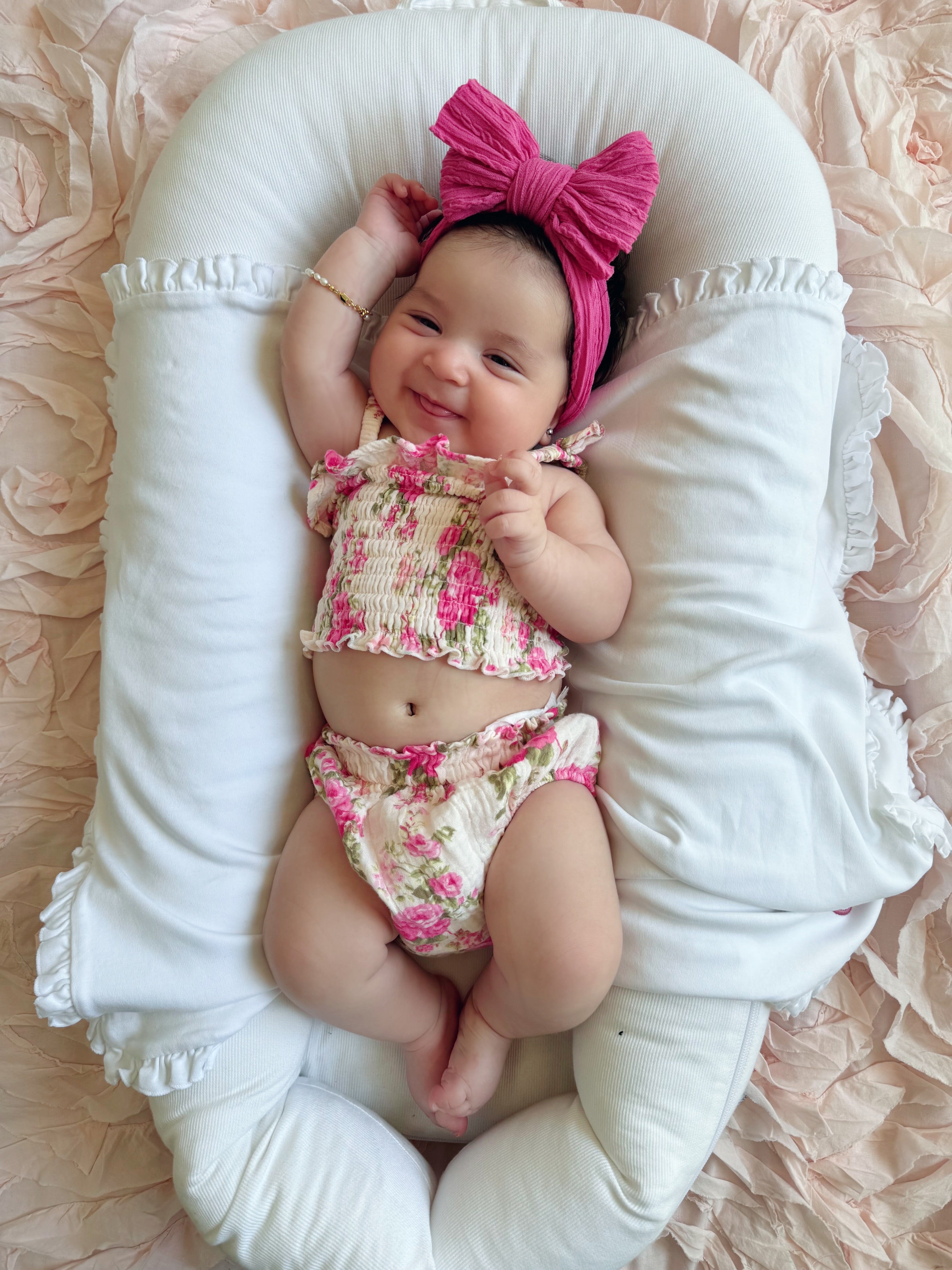 Smiling baby girl in floral outfit with pink bow, lying on soft background, radiating joy and cuteness.