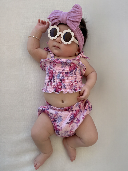 Baby girl in floral outfit with sunglasses and a headband, lying on a light background.